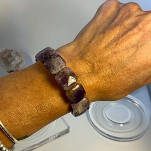 Puede incluir: Un brazalete con grandes piedras de amatista de forma irregular. Las piedras, en tonos morados y blancos, están ensartadas juntas. El brazalete grueso se muestra en una muñeca, destacando las variaciones naturales de las piedras preciosas.