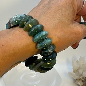 Puede incluir: Un brazalete de piedras preciosas verdes y turquesas. El brazalete está hecho de cuentas de forma ovalada, ensartadas. Las cuentas tienen un acabado pulido y varían en color de verde oscuro a turquesa. El brazalete se lleva en la muñeca.