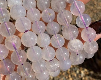 Könnte beinhalten: Eine Strähne aus irisierenden weißen Quarzperlen mit einem dezenten violetten Farbton. Die Perlen sind rund und glatt und auf einer durchsichtigen Gummikordel aufgefädelt.