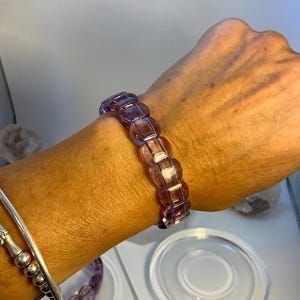 Puede incluir: Una pulsera de color lavanda con cuentas rectangulares se lleva en la muñeca. La pulsera está hecha de piedras translúcidas y pulidas. Se ven otras joyas en la muñeca, incluyendo un brazalete de plata y una pulsera con cuentas redondas. La pulsera es una joya.