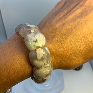 Puede incluir: Un brazalete grueso con grandes cuentas redondeadas en tonos grises, blancos y verde pálido. Las cuentas tienen un aspecto veteado y están ensartadas para formar el brazalete. El brazalete se lleva en la muñeca.