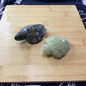 May include: Two stone turtle figurines. One is a speckled grey and black, the other is a pale green. Both are carved in a turtle shape and sit on a light brown wooden surface. The turtles are likely decorative items.