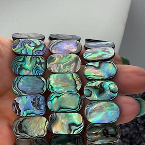 Puede incluir: Pulsera de concha de abulón con iridiscencias azules, verdes y púrpuras. Las cuentas rectangulares están ensartadas, mostrando los patrones y colores naturales de la concha. La pulsera se sostiene sobre un fondo neutro.