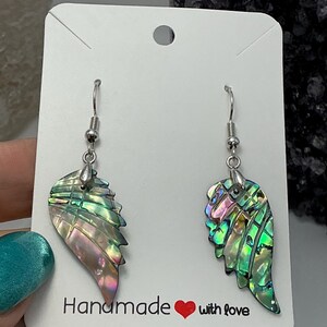 Puede incluir: Un par de pendientes iridiscentes en forma de ala con ganchos plateados. Los pendientes muestran tonos verdes, rosas y morados. Se presentan en una tarjeta blanca con el texto "Handmade with love".