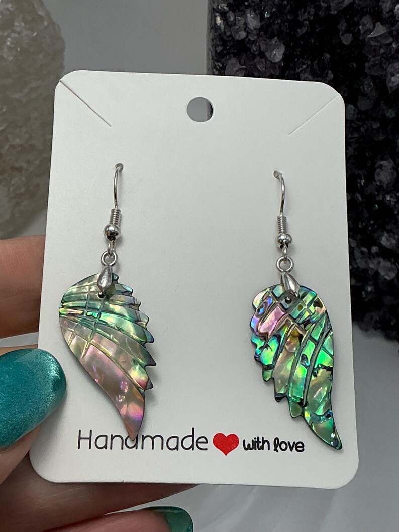 Puede incluir: Un par de pendientes iridiscentes en forma de ala con ganchos plateados. Los pendientes muestran tonos verdes, rosas y morados. Se presentan en una tarjeta blanca con el texto "Handmade with love".