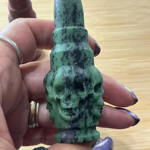 May include: A green and black stone sculpture of a skull with a cylindrical top. The sculpture has a rough texture and is held in a person's hand. Another similar sculpture is visible in the background.