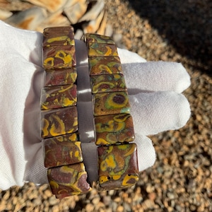 May include: Two matching bracelets with rectangular, patterned beads. The beads feature a mix of brown, yellow, and green colors. The bracelets are displayed on a white surface.