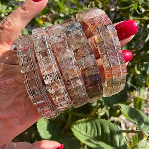 May include: Five clear quartz bracelets with inclusions of black, brown, and red. The bracelets are stacked on top of each other and are made of square-shaped beads.