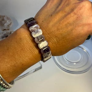 Puede incluir: Un brazalete hecho de piedras de amatista rectangulares en tonos morado, blanco y negro. El brazalete se lleva en la muñeca, mostrando las variaciones naturales de las piedras preciosas. El brazalete es una joya.