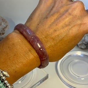 Puede incluir: Un brazalete translúcido de color rojo púrpura compuesto por cuentas rectangulares. El brazalete se lleva en la muñeca, con un anillo plateado visible en un dedo. También es visible un brazalete de cuentas turquesas.