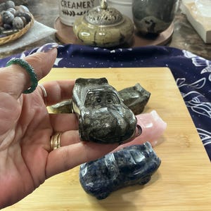 May include: A collection of carved stone car figurines in various colors, including gray, pink, and blue. The cars are displayed on a wooden surface, with a hand holding one of the gray cars. Other decorative items are in the background.