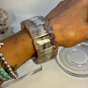 Puede incluir: Un brazalete ancho hecho de piedras de ágata pulidas. El brazalete presenta piedras rectangulares y bandeada en tonos grises, blancos y marrón rojizo. Las piedras están unidas para formar una pieza de joyería llamativa.