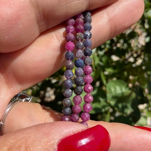 May include: A strand of faceted gemstone beads in shades of pink, purple, and blue. The beads are round and have a slightly irregular shape.