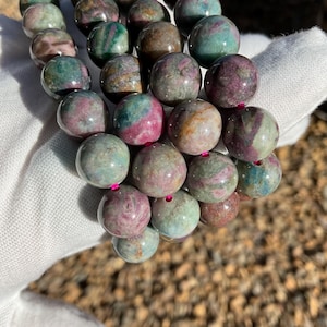 May include: A multi-strand bracelet made of round, green, pink, and brown gemstone beads. The beads have a swirling pattern and are polished to a smooth finish.