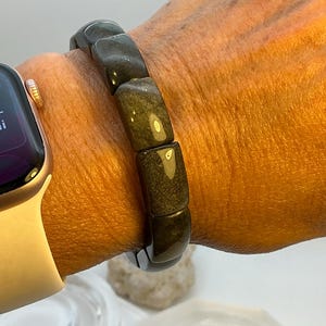 May include: A dark, rectangular bead bracelet with a gold sheen. The bracelet is worn on a wrist next to a gold smartwatch with a white band. The beads are smooth and polished, reflecting light.