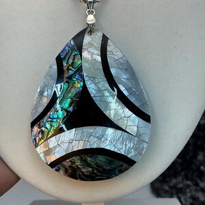 May include: A teardrop-shaped pendant with a silver chain. The pendant features a mosaic design with iridescent abalone shell, black, and white sections. The abalone shell displays a range of colors, including green, blue, and purple.