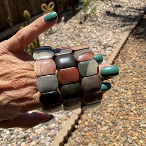 May include: Three stacked bracelets made of various shades of brown, grey, black, and green stone. The stones are rectangular and polished.