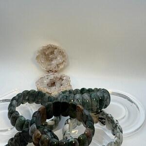 Puede incluir: Se muestran varias pulseras de piedras preciosas en tonos verdes, marrones y blancos. Las pulseras están hechas de cuentas rectangulares pulidas. Una geoda está en el fondo. Las pulseras están sobre una superficie redonda transparente.