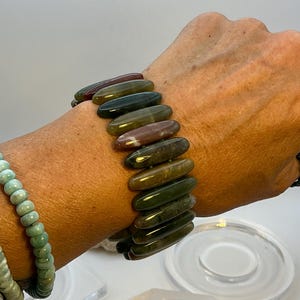 Op de afbeelding: Een armband met langwerpige, gepolijste stenen in groen, bruin en bordeaux. De armband wordt om de pols gedragen en toont de natuurlijke kleurvariaties. Een tweede armband met groene kralen is ook zichtbaar.