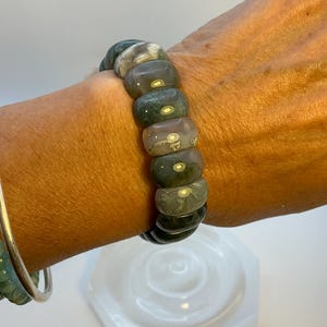 Puede incluir: Una pulsera de cuentas con cuentas grandes y redondeadas en tonos verdes, grises y crema. La pulsera se lleva en la muñeca, mostrando los patrones y texturas de la piedra natural. También se ve una pulsera plateada.