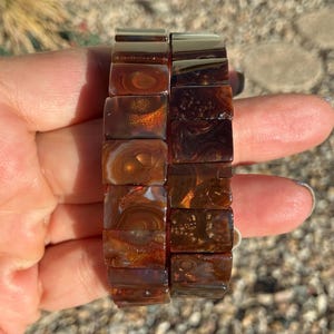 May include: Two stretch bracelets composed of rectangular, polished brown agate beads. The beads feature natural, swirling patterns in shades of deep amber, mahogany, and dark chocolate, with translucent areas that catch the light to reveal internal banding.