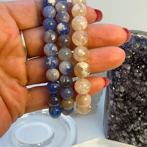 Puede incluir: Tres pulseras de cuentas. Una pulsera presenta cuentas azules y blancas, otra tiene cuentas grises y blancas, y la tercera tiene cuentas color melocotón. Las pulseras están hechas de piedras redondas y pulidas.