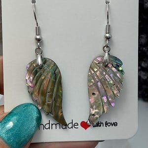Puede incluir: Un par de pendientes iridiscentes en forma de ala con ganchos plateados. Los pendientes est&aacute;n hechos de un material similar a una concha con tonos verdes, rosas y morados. La tarjeta detr&aacute;s de los pendientes dice "handmade with love".