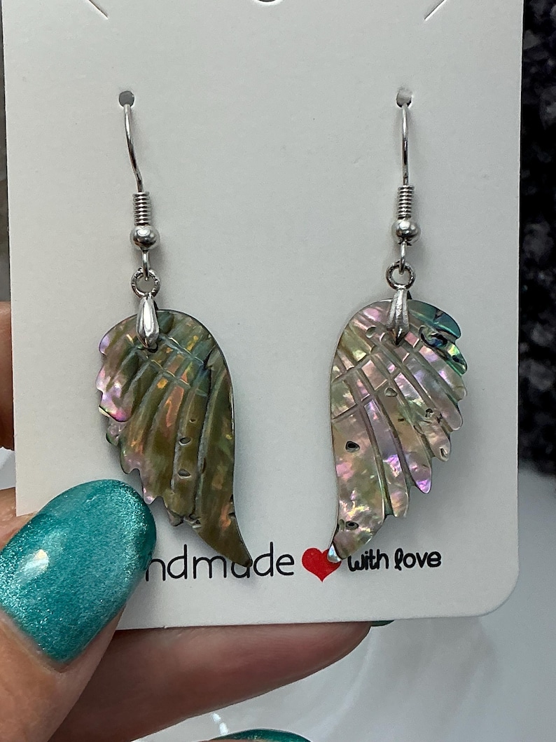 Puede incluir: Un par de pendientes iridiscentes en forma de ala con ganchos plateados. Los pendientes est&aacute;n hechos de un material similar a una concha con tonos verdes, rosas y morados. La tarjeta detr&aacute;s de los pendientes dice "handmade with love".