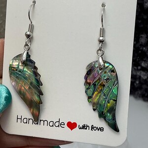 Puede incluir: Un par de pendientes con forma de ala hechos con concha de abul&oacute;n iridiscente. Los pendientes tienen ganchos plateados y se exhiben en una tarjeta blanca con el texto "Handmade with love" y un coraz&oacute;n rojo.