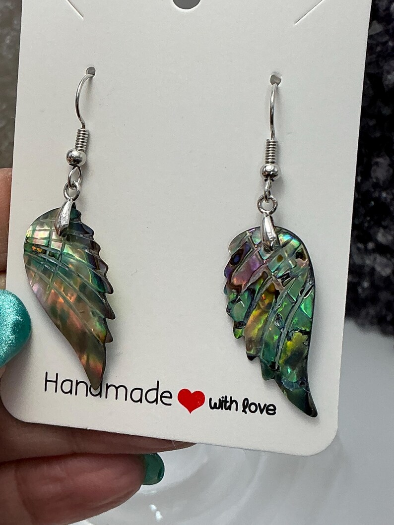 Puede incluir: Un par de pendientes con forma de ala hechos con concha de abul&oacute;n iridiscente. Los pendientes tienen ganchos plateados y se exhiben en una tarjeta blanca con el texto "Handmade with love" y un coraz&oacute;n rojo.