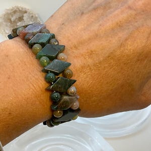 Puede incluir: Una pulsera de piedras preciosas con una mezcla de cuentas verdes, marrones y beiges y piedras en forma de diamante. La pulsera se lleva en la muñeca, mostrando los colores y texturas naturales de las piedras. Es una joya.
