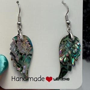 Puede incluir: Un par de pendientes hechos a mano con forma de ala. Los pendientes presentan conchas de abul&oacute;n iridiscentes en tonos verdes, azules y morados. El texto "Handmade with love" est&aacute; impreso en la tarjeta blanca. Los pendientes tienen ganchos plateados.