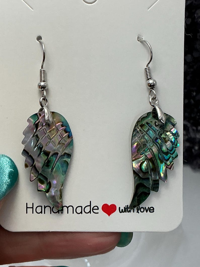 Puede incluir: Un par de pendientes hechos a mano con forma de ala. Los pendientes presentan conchas de abul&oacute;n iridiscentes en tonos verdes, azules y morados. El texto "Handmade with love" est&aacute; impreso en la tarjeta blanca. Los pendientes tienen ganchos plateados.