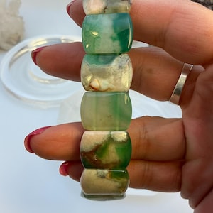 May include: A stacked bracelet composed of six barrel-shaped beads. The beads are a mix of green, white, and brown, with a polished, glossy finish. The bracelet is held in a hand with red nail polish and a silver ring.