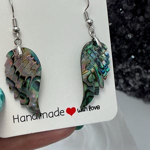Puede incluir: Pendientes hechos a mano con colgantes de ala de concha de abul&oacute;n iridiscente. Los pendientes tienen ganchos plateados y se exhiben en una tarjeta blanca con el texto "Handmade with love". Las alas muestran tonos de verde, azul y morado.