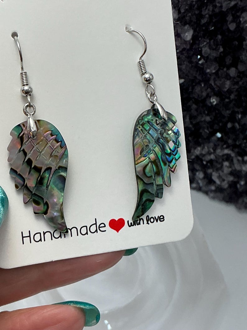 Puede incluir: Pendientes hechos a mano con colgantes de ala de concha de abul&oacute;n iridiscente. Los pendientes tienen ganchos plateados y se exhiben en una tarjeta blanca con el texto "Handmade with love". Las alas muestran tonos de verde, azul y morado.