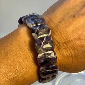 Puede incluir: Una pulsera elástica con grandes cuentas de amatista pulidas. Las cuentas son de color púrpura intenso con vetas blancas y púrpuras más claras. La pulsera se lleva en la muñeca, mostrando la belleza y los patrones únicos de la piedra natural.