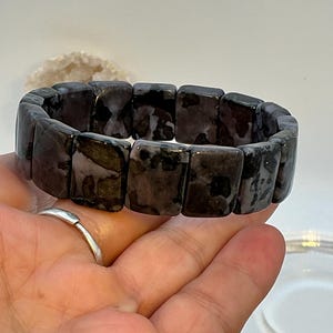 May include: A dark gray and black gemstone bracelet with rectangular beads. The bracelet is held by a hand with a silver ring. The beads have a mottled pattern and a polished finish. The background is a light, neutral color.