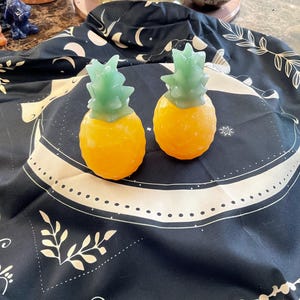 Op de afbeelding: Twee decoratieve objecten in de vorm van ananassen, met gele lichamen en groene toppen. Ze liggen op een zwarte doek met een wit hemels patroon. De objecten zijn waarschijnlijk gemaakt van een vast materiaal, mogelijk zeep of was.