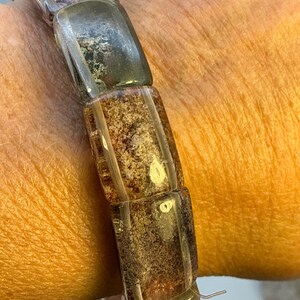May include: A stretch bracelet composed of rectangular, translucent beads in shades of amber, brown, and dark gray. Each bead features a unique, textured, and mottled internal pattern, creating a varied appearance across the length of the bracelet.