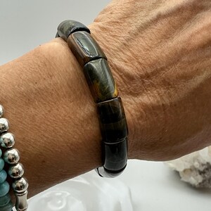 May include: A dark, rectangular bead bracelet with a mix of dark green, brown, and gold tones. The bracelet is worn on a wrist, showcasing the polished, cylindrical beads. The bracelet is a unique piece of jewelry.