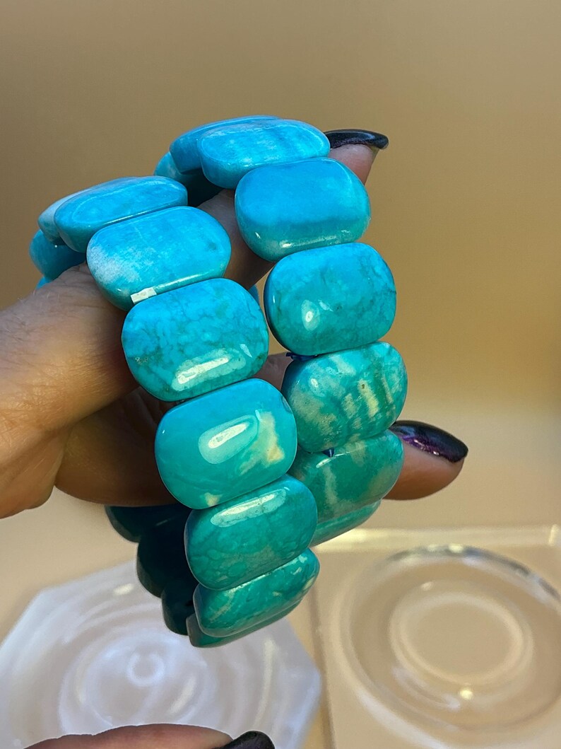May include: A stretch bracelet composed of smooth, rectangular turquoise-colored beads with natural veining and variations in shade. The beads are arranged in a continuous loop, showcasing a polished finish and a vibrant, saturated blue-green hue.