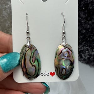 May include: Oval-shaped abalone shell earrings with iridescent colors of green, purple, and blue. The earrings hang from silver-colored hooks. The earrings are displayed on a white card with the word "made" and a red heart.
