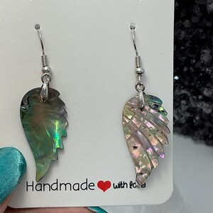 Puede incluir: Un par de pendientes iridiscentes de concha de abul&oacute;n con forma de ala. Los pendientes cuelgan de ganchos plateados. Se exhiben en una tarjeta blanca con la frase "Handmade with love".