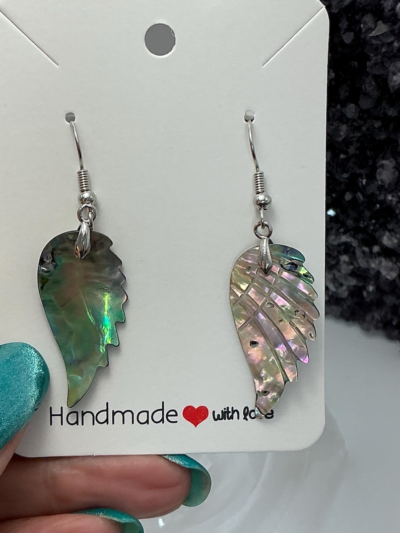Puede incluir: Un par de pendientes iridiscentes de concha de abul&oacute;n con forma de ala. Los pendientes cuelgan de ganchos plateados. Se exhiben en una tarjeta blanca con la frase "Handmade with love".