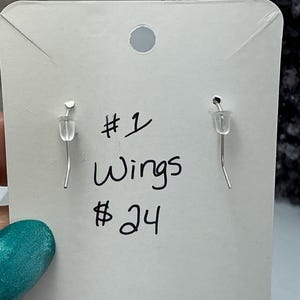 Puede incluir: Pendientes plateados con dise&ntilde;o de gancho, exhibidos en una tarjeta blanca. La tarjeta tiene texto escrito a mano que dice "#1 Wings". El fondo est&aacute; desenfocado.