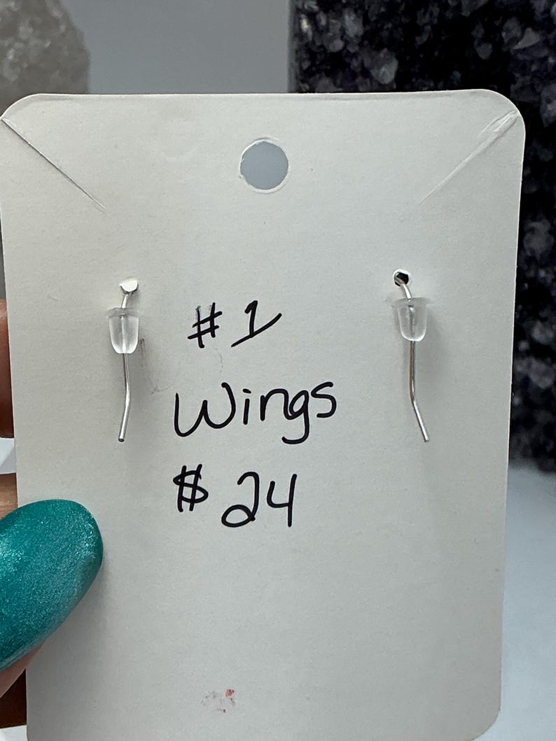Puede incluir: Pendientes plateados con dise&ntilde;o de gancho, exhibidos en una tarjeta blanca. La tarjeta tiene texto escrito a mano que dice "#1 Wings". El fondo est&aacute; desenfocado.