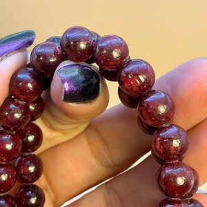 May include: A strand of polished, deep red garnet beads arranged in a circular formation. Each spherical bead features a smooth, reflective surface with natural internal inclusions and variations in color intensity throughout the gemstone strand.