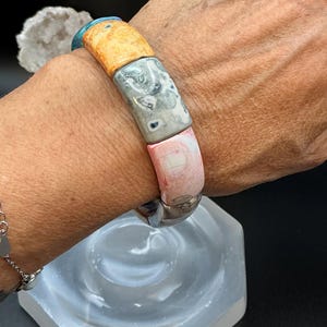 Op de afbeelding: Een elastische armband bestaande uit rechthoekige kralen in diverse kleuren en patronen, waaronder turkoois, geel, grijs met donkere spikkels en roze met witte marmering, gerangschikt in een reeks rond de pols.