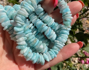 Puede incluir: Un collar hecho de cuentas irregulares de color azul claro y blanco. Las cuentas están ensartadas, formando una forma circular. El collar se sostiene en una mano, con un fondo natural de follaje verde y flores rosas.